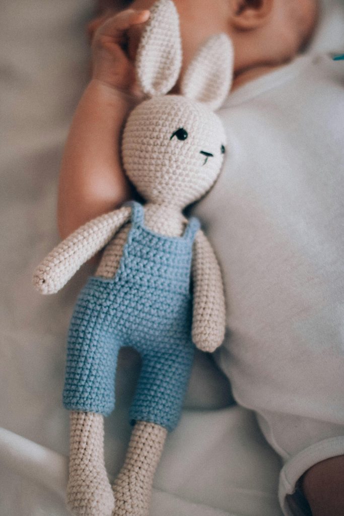 A cute baby lies next to a handmade crochet bunny doll in soft lighting.